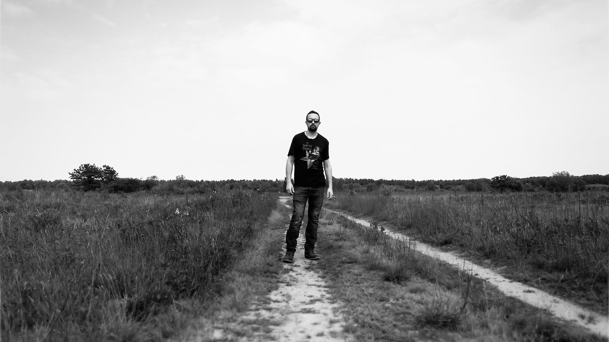 Black-and-white environmental portrait of Thomas Stirling standing on a sandy path amid wide-open coastal barrens beneath a vast sky